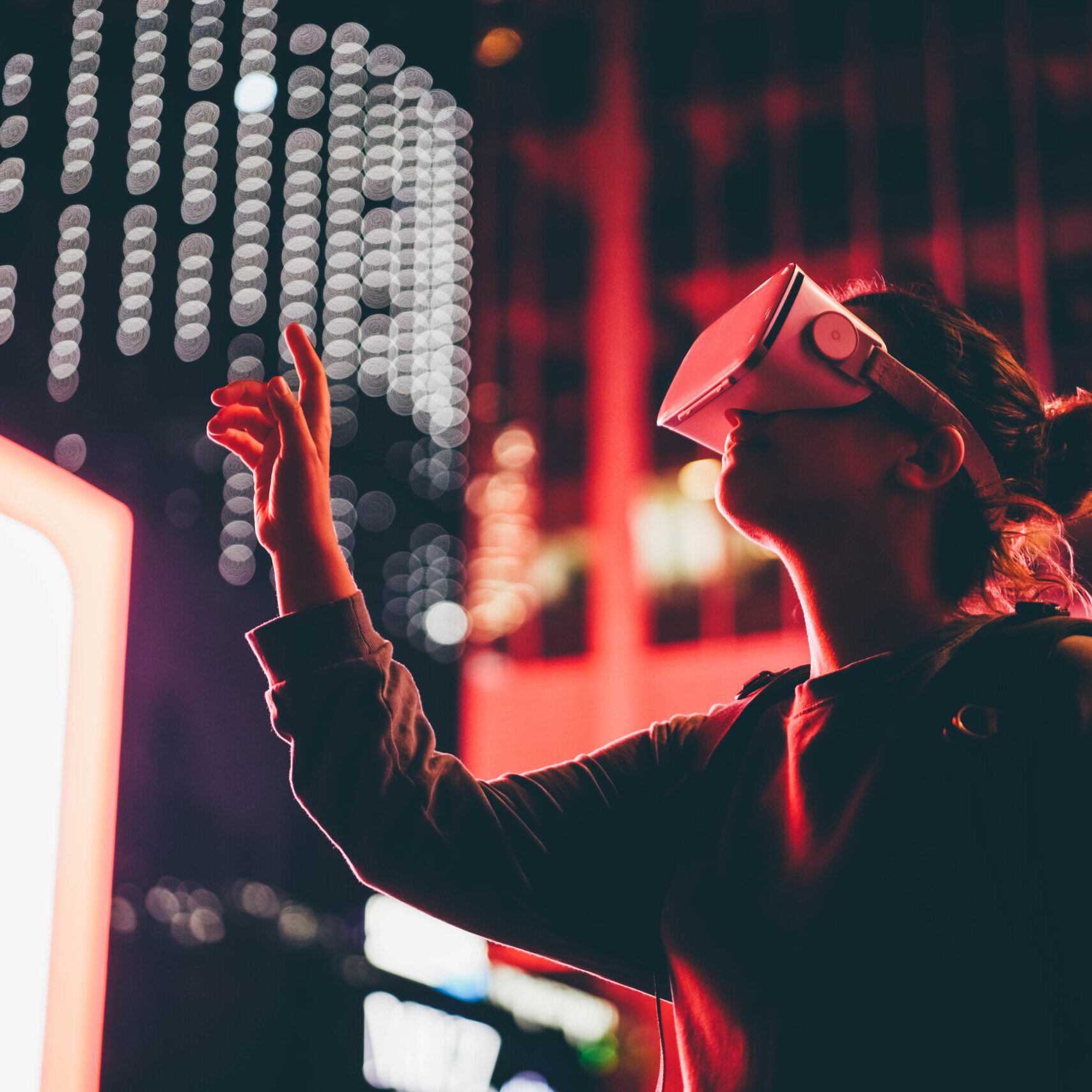 Young woman in virtual reality glasses with backpack looks around and moves hands standing against big modern financial center buildings in late evening.